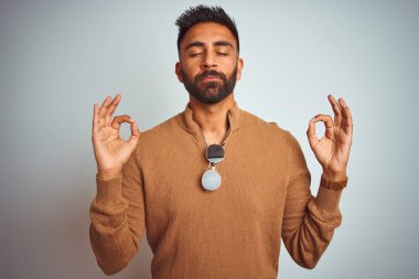 Zarif süveter ve güneş gözlüğü takan Hintli adam izole edilmiş beyaz arka planda durup rahatlıyor ve parmaklarıyla meditasyon hareketi yaparken gözleri kapalı gülümsüyor. Yoga konsepti.
