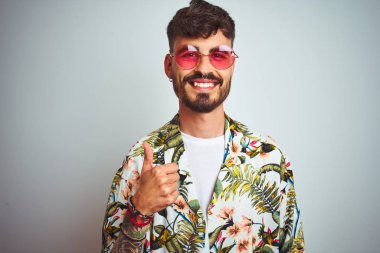 Tatilde dövmeli, güneş gözlüğü takan, izole edilmiş beyaz arka planda mutlu bir el hareketi yapan bir adam. Kameraya bakıp başarıyı gösteren ifade onaylanıyor.