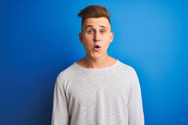 İzole edilmiş mavi arka plan üzerinde duran beyaz tişörtlü genç, yakışıklı bir adam şaşkınlık dolu bir yüz ifadesinden korkuyor ve heyecanlanıyor.
