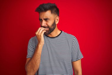 İzole edilmiş kırmızı arka plan üzerinde denizci çizgili tişört giyen genç, yakışıklı Hintli adam elleri ağız yiyen tırnaklarla gergin ve stresli görünüyor. Endişe sorunu.
