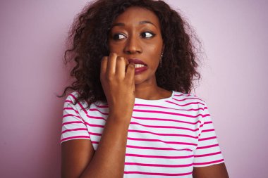 İzole edilmiş pembe arka planda duran çizgili tişört giyen genç Afro-Amerikalı kadın elleri ağız yiyen tırnaklarla gergin ve stresli görünüyor. Endişe sorunu.