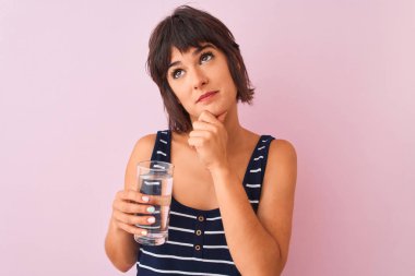 Genç ve güzel bir kadın bir bardak su tutuyor pembe arka planda duruyor ciddi bir yüz soru soruyor, kafası çok karışık bir fikir.