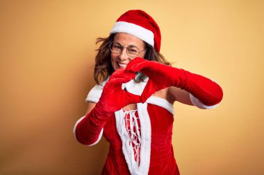 Orta yaşlı, güzel bir kadın, izole edilmiş sarı arka planda Noel Baba kostümü giyiyor. Sevgiyle gülümsüyor, elleriyle kalp sembolü ve şekli gösteriyor. Romantik konsept.