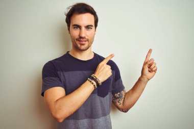 Izole beyaz arka plan üzerinde rahat t-shirt giyen dövme ile genç yakışıklı adam gülümseyerek ve yan iki el ve parmakları ile işaret kamera bakıyor.