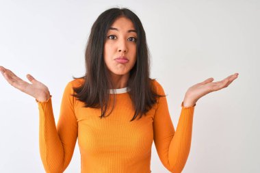 Turuncu tişört giyen genç, güzel Çinli bir kadın izole edilmiş beyaz arka plan üzerinde duruyor. Kolları ve elleri havada, kafası karışmış bir ifade takınıyor. Şüphe kavramı.