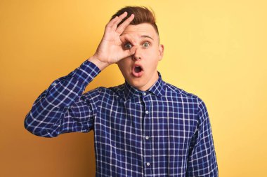 Sarı arka plan üzerinde duran sıradan tişörtlü genç yakışıklı adam şaşırmış yüz ifadesiyle şaşırmış ve parmaklarından içeri bakıyor. İnançsız ifade.