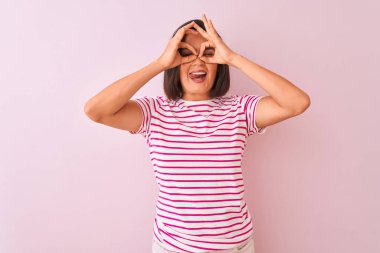 İzole edilmiş pembe arka plan üzerinde duran çizgili tişört giyen genç ve güzel bir kadın dürbünün dili dışarı çıkarması, gözlerin parmaklarından içeri bakması gibi güzel bir jest yapıyor. Çılgın ifade.