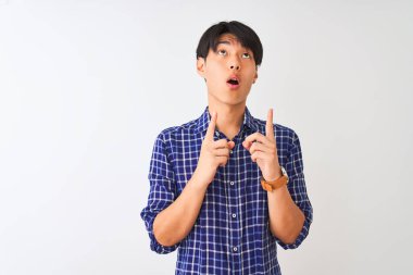 İzole edilmiş beyaz arka plan üzerinde duran gündelik mavi gömlekli genç Çinli adam şaşırmış ve şaşırmış bir şekilde yukarı bakıyor ve parmaklarıyla işaret ediyor ve kollarını kaldırıyor..