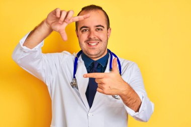 Ceket ve steteskop giyen genç doktor izole edilmiş sarı arka planda durup gülümsüyor ve mutlu yüzlü elleri ve parmakları olan bir çerçeve yapıyor. Yaratıcılık ve fotoğrafçılık kavramı.
