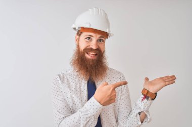 İzole edilmiş beyaz arka plan üzerinde güvenlik kaskı takan genç kızıl saçlı mimar kameraya hayret ediyor ve gülümsüyor..