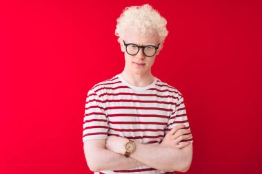 İzole edilmiş kırmızı arka plan üzerinde çizgili tişört ve gözlük takan genç, sarışın adam şüpheci ve gergin, yüzünde çapraz kollu tasvip etmeyen bir ifade var. Negatif kişi.