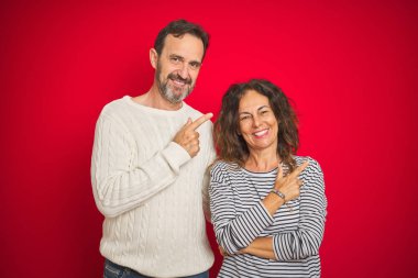 Güzel bir orta yaşlı çift, izole edilmiş kırmızı arka plan üzerine kışlık süveter giyer. Neşeli, yüzünde mutlu ve doğal bir ifadeyle, elleri ve parmaklarıyla yüzünü gösteren bir gülümseme.