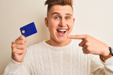 Genç ve yakışıklı bir adam kredi kartını soyutlanmış beyaz bir arkaplanda tutuyor. Elle ve parmakla göstererek çok mutlu.