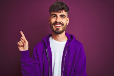 İzole edilmiş sarı arka planda duran çizgili polo giyen genç bir adam yüzünde kocaman bir gülümsemeyle kameraya el ve parmak ile bakıyor..