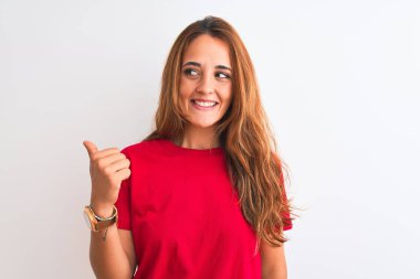 Kızıl saçlı, kırmızı tişörtlü genç bir kadın. İzole edilmiş beyaz bir arka plan üzerinde gülümsüyor. Gülümsüyor ve başparmağıyla yan tarafı işaret ediyor..