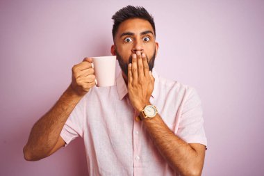 Genç Hintli bir adam gömleğini giyip kahve içerken izole edilmiş pembe arka planda duruyor. Ağzını kapatıyor. Yanlışlıkla utancından ağzı sarsılıyor.