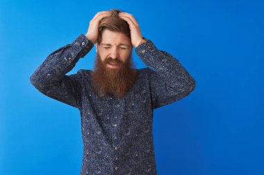 Çiçekli yaz gömleği giyen genç kızıl saçlı adam izole edilmiş mavi arka planda duruyor. Ağrı ve migren yüzünden baş ağrısı ve stresten muzdarip. Ellerini başının üstüne koy..