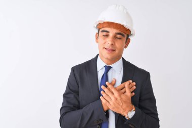 İzole edilmiş beyaz arka plan üzerinde kask ve takım elbise giyen genç, yakışıklı mimar elleri göğsünde, gözleri kapalı ve yüzünde minnet dolu bir jest ile gülümsüyor. Sağlık kavramı.