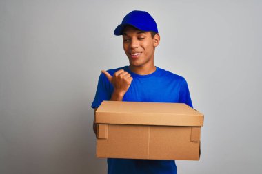 Genç yakışıklı arap teslimat adam izole beyaz arka plan üzerinde duran bir kutu tutan işaret ve mutlu yüzü gülümseyerek yan başparmak ile gösteren