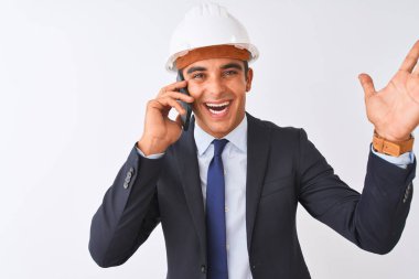 Miğfer takan genç mimar, kapalı beyaz arka planda akıllı telefondan konuşuyor. Çok mutlu ve heyecanlı. Kazanan ifade zaferi kutluyor.