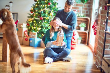 Mutlu ve kendinden emin gülümseyen köpeği olan genç ve güzel bir çift. Kadının gözlerini kapayan bir adam Noel ağacının etrafında hediyeyle dolaşır.