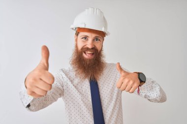 İzole edilmiş beyaz arka plan üzerinde güvenlik kaskı takan genç kızıl saçlı mimar el hareketini onaylıyor, gülümsüyor ve başarı için mutlu. Kazanan jest.
