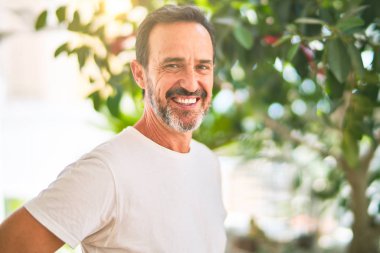 Orta yaşlı yakışıklı bir adam terasta dikilmiş gülümseyerek günlük tişört giyiyor.