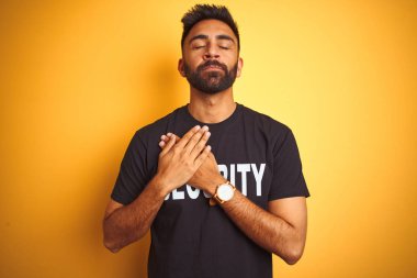 Arap Hindistanlı İspanyol güvenlik üniforması giyen izole edilmiş sarı arka planda elleri göğsünde, gözleri kapalı ve yüzünde minnet dolu bir jest ile gezen adam. Sağlık kavramı.