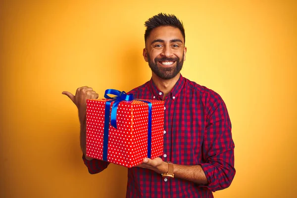Arab indian hispanic man holding birthday gift standing over isolated yellow background pointing and showing with thumb up to the side with happy face smiling
