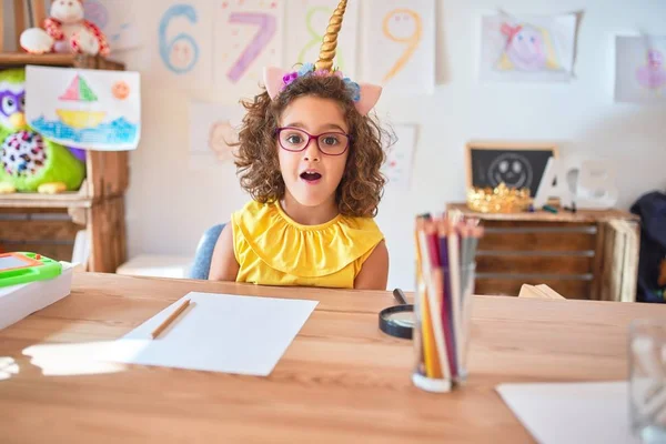 Gözlüklü ve tek boynuzlu at diademi takmış güzel bir çocuk anaokulunda masasında korkmuş ve şok olmuş bir şekilde oturuyor..