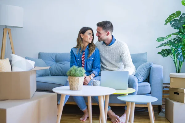 Genç güzel çift karton kutular etrafında yeni evde dizüstü bilgisayar kullanarak kahve kanepede içme fincan oturan