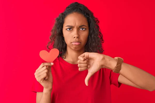 年轻的巴西女人拿着纸心站在孤立的红色背景上 满脸怒容 负面的迹象表明她不喜欢大拇指朝下 拒绝的概念 — 图库照片