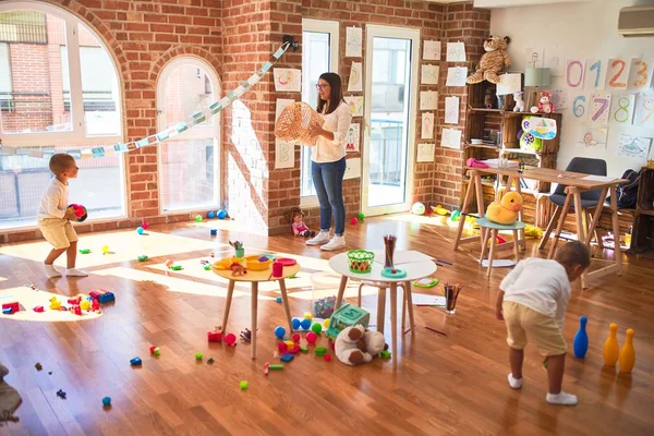 Anaokulunda bir sürü oyuncağın etrafında top ve hasır sepet kullanarak basketbol oynayan güzel öğretmenler ve yeni yürümeye başlayan çocuklar.