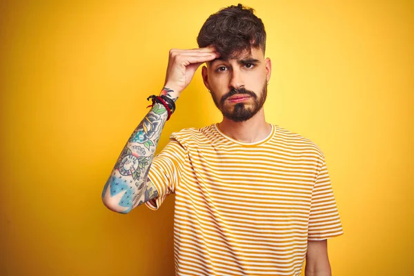 Dövmeli, çizgili tişörtlü genç adam izole edilmiş sarı arka planda duruyor. Alnındaki sorun yüzünden endişeli ve stresli. Kriz konusunda endişeli.