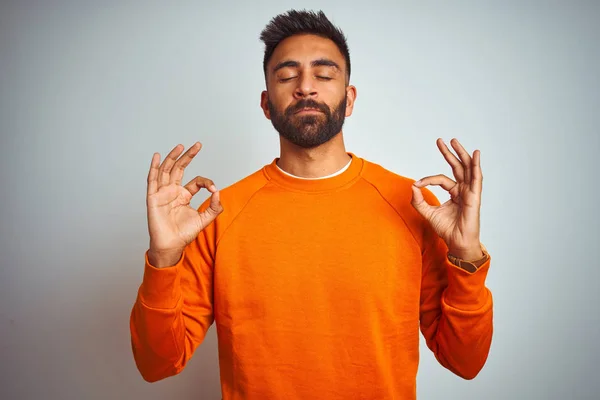 Young indian man wearing orange sweater over isolated white background relax and smiling with eyes closed doing meditation gesture with fingers. Yoga concept.