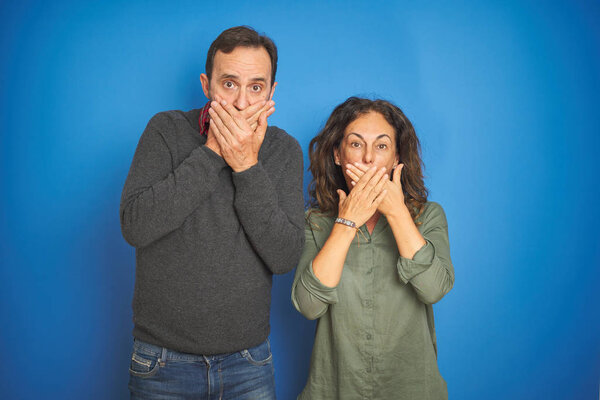 Beautiful middle age couple together standing over isolated blue background shocked covering mouth with hands for mistake. Secret concept.