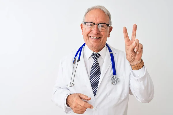 独立した白い背景の上に立って聴診器を身に着けているシニア白髪の医者の男は 指が勝利のサインを行うことを示すカメラを見て微笑んだ 第二番 — ストック写真