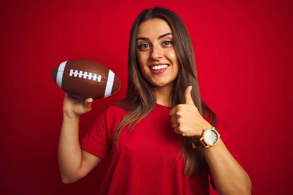 年轻漂亮的女运动员抱着足球在孤立的红色背景上开心地笑着做着好的手势 用手指竖起大拇指 很好的标志 — 图库照片