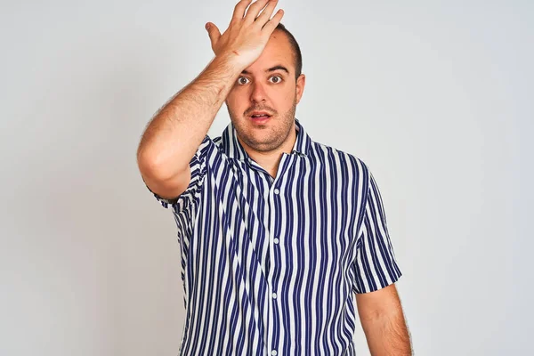 Young man wearing blue striped shirt standing over isolated white background surprised with hand on head for mistake, remember error. Forgot, bad memory concept.