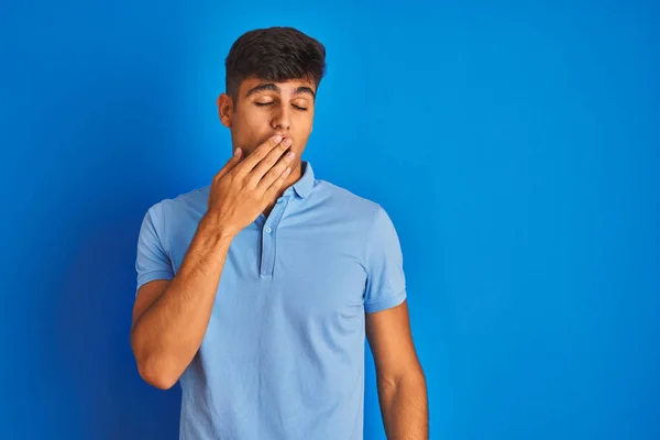 Young indian man wearing casual polo standing over isolated blue background bored yawning tired covering mouth with hand. Restless and sleepiness.