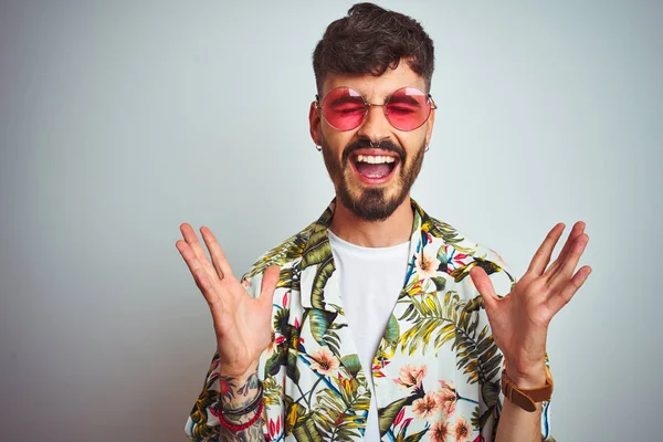 Tatilde dövmeli bir adam, güneş gözlüğü takıyor, izole edilmiş beyaz arka planda çılgın ve başarıyı kutluyor, kolları havada ve gözleri kapalı, heyecanlı bir şekilde bağırıyor. Kazanan konsept