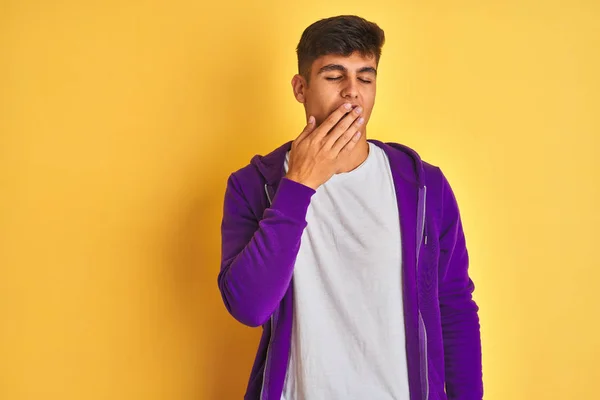 Young indian man wearing purple sweatshirt standing over isolated yellow background bored yawning tired covering mouth with hand. Restless and sleepiness.
