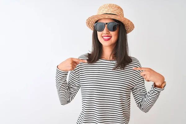Chinese woman wearing striped t-shirt hat sunglasses standing over isolated white background looking confident with smile on face, pointing oneself with fingers proud and happy.