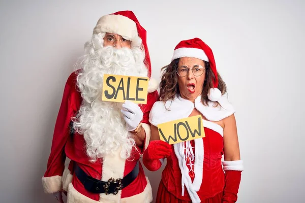 Couple wearing Santa costume holding wow and sale banner over isolated white background In shock face, looking skeptical and sarcastic, surprised with open mouth
