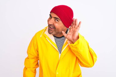 Yağmurluk ve yünlü şapka giyen orta yaşlı bir adam izole edilmiş beyaz arka planda dikiliyor. Kulaktan kulağa gülümsüyor ve dedikodulara ve dedikodulara kulak veriyor. Sağırlık kavramı.
