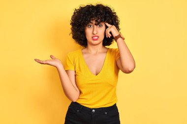 Sarı arka plan üzerinde duran kıvırcık saçlı genç Arap kadın avuç içi açık fotokopi alanı ve alnını işaret ederek kafasını karıştırıyor ve rahatsız ediyor. Bunu bir düşün..