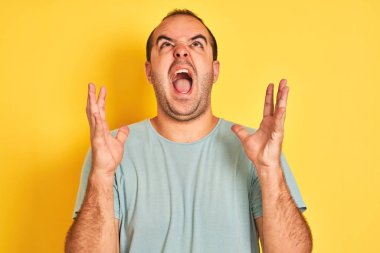 Yeşil tişört giyen genç adam izole edilmiş sarı arka planda çılgın deli bağırıyor ve agresif bir ifade ve kollarla bağırıyor. Hayal kırıklığı konsepti.
