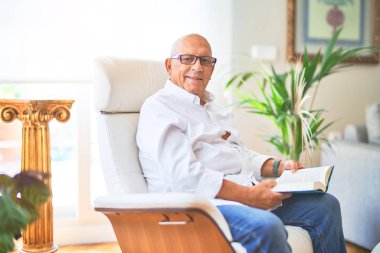 Senior handsome man smiling happy and confident. Sitting on the armchair relaxed reading book at home