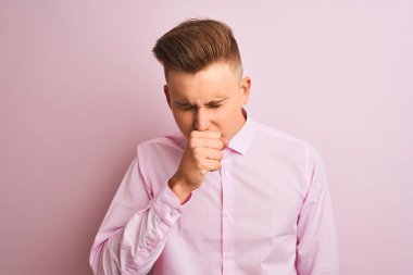 Soyutlanmış pembe arka plan üzerinde duran şık gömlekli genç yakışıklı işadamı kendini iyi hissetmiyor ve soğuk algınlığı veya bronşitin belirtisi olarak öksürüyor. Sağlık hizmetleri kavramı.