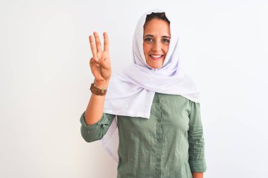 Geleneksel Müslüman tesettürlü genç, güzel bir Arap kadını izole edilmiş arka planda üç numaralı parmaklarıyla işaret ederken kendinden emin ve mutlu bir şekilde gülümsüyor..